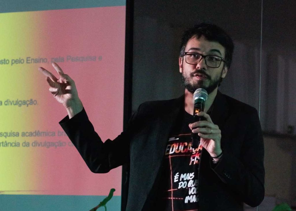 Leandro Rincon, coordenador de pesquisa da Faculdade EnsinE, vestindo um blazer preto e seguranod um microfone durante palestra.